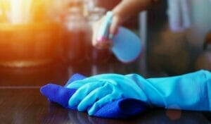 Hands of an individual cleaning: one hand wearing a glove holding a cloth, and the other hand without a glove holding a cleaning spray.