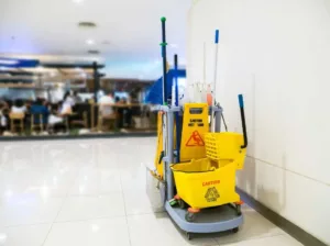 Professional cleaning team with a yellow cart filled with cleaning products and a "caution wet floor" sign, situated in a clean, white corridor with shiny tiles, with a blurred background of a restaurant resembling an airport setting.