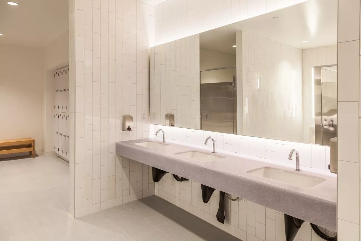 A bathroom that resembles a gym or office setting, featuring a sink with a backlit mirror, lockers in the background, and a stainless steel door possibly leading to showers or toilets.