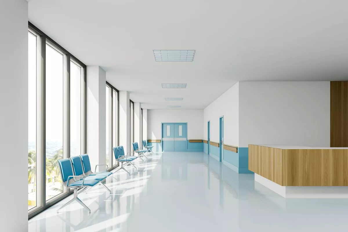 A modern hospital corridor featuring a window on the left with blue chairs, a shiny floor, and a counter on the right.
